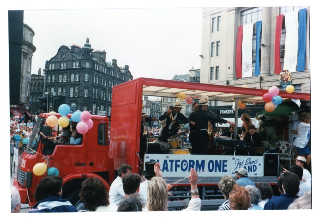 A jazz band performs from inside a decorated red lorry to a crowded pavement audience as the lorry turns up Lothian Road from Princes Street.