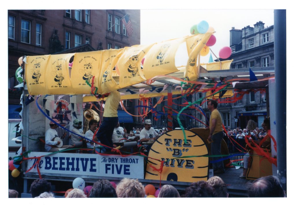 A group of musicians perform from within an open-sided lorry decorated with yellow flags and a 'B' hive as it goes along the foot of Lothian Road.