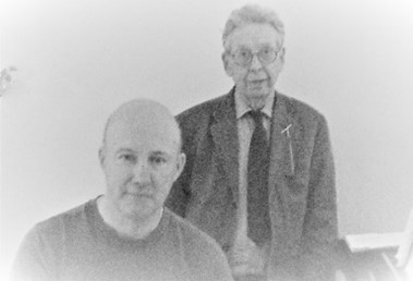 Black and white portrait of two men, one in a t shirt and the other in glasses and jacket and tie.