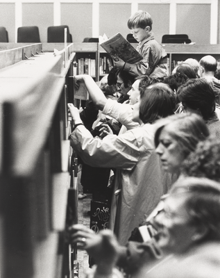A boy is sitting on someone's shoulders reading a book amidst a crowd of people searching through bookshelves.