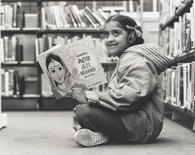 A girl sits cross-legged on the floor of a library reading a picture book.
