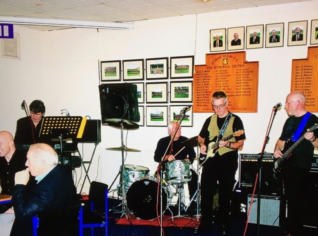 A band of four men playing instruments at a rugby club venue.