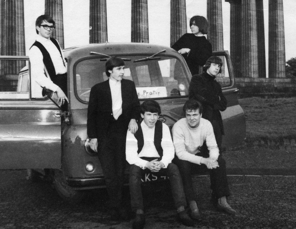 Six men are posed for a group portrait in and around an old van parked in front of the National Monument on Calton Hill.