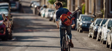 A young man cycles down a road lined with parked cars with a guitar strapped to his back.