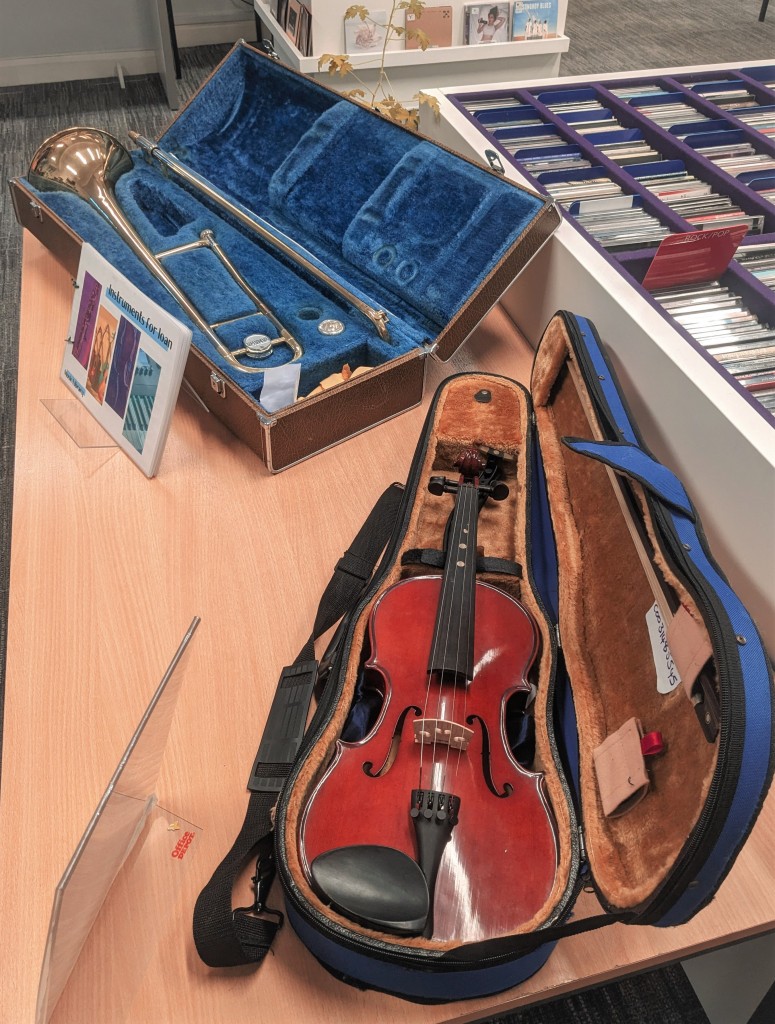 A trombone and a violin both in their open cases are displayed on a table beside a large rack of CDs.