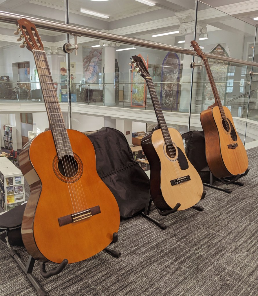 Three guitars are displayed on stands on the floor of a mezzanine in front of a glass barrier overlooking the floor below.