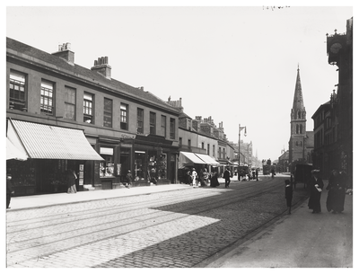 An historical view looking along the High Street at Portobello with shops lining the far side of the street and people walking on either side.