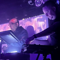 Two men stand on a stage lit with purple light at electronic keyboards.