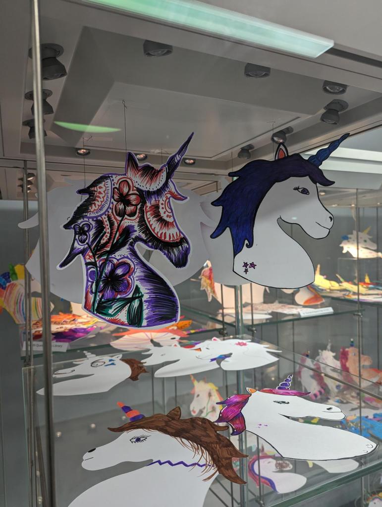 Lots of decorated unicorn heads are displayed on different levels of a glass cabinet with two hanging in the foreground.