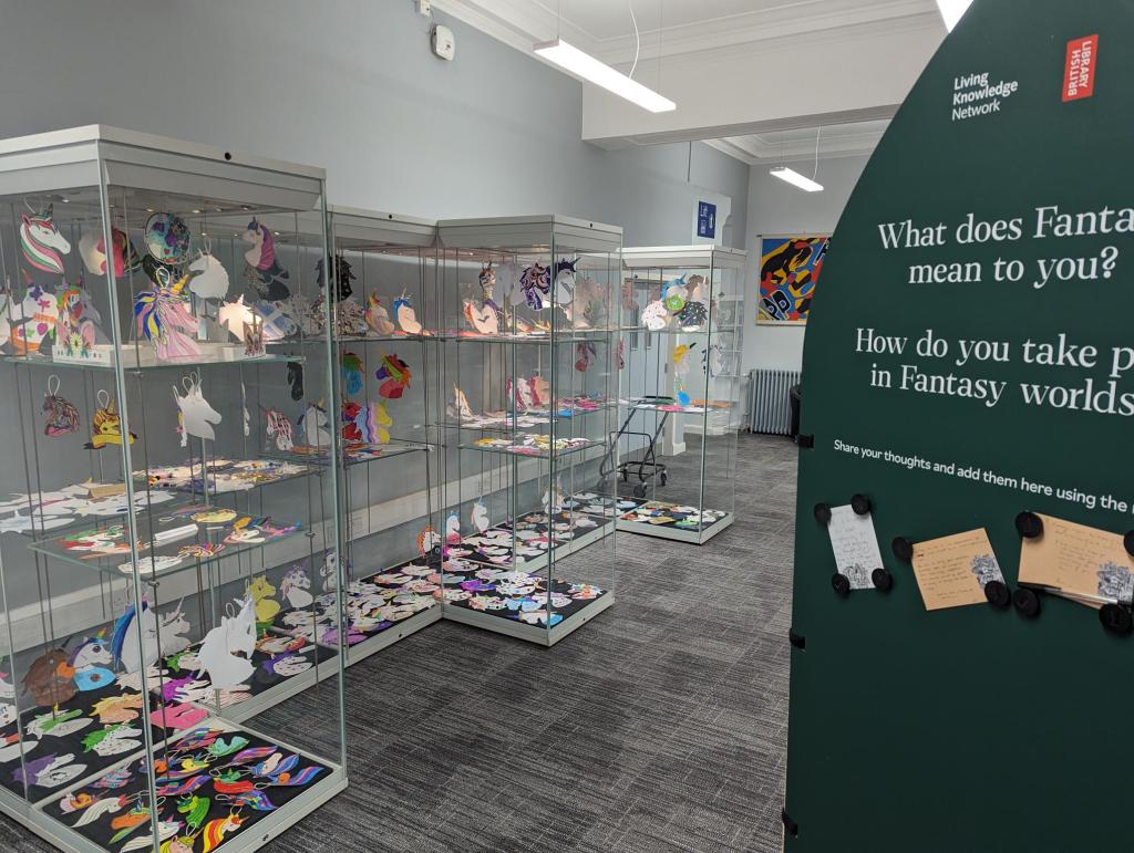 Lots of decorated paper unicorns are displayed in a large shelved glass cabinet next to a green display board.