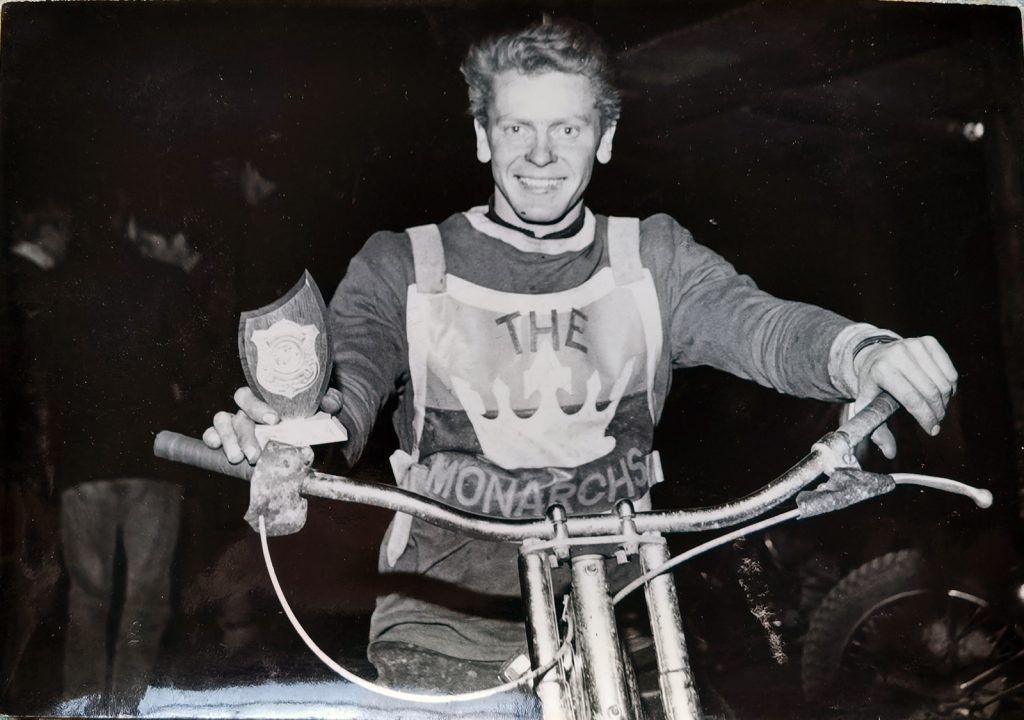 A man sits on a bike holding the handlebars and a trophy and is wearing a sign saying, "The Monarchs" over his t-shirt.
