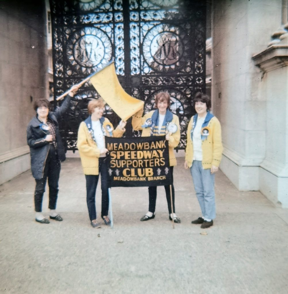 A group of four women, three in yellow jackets stand in front of a large, ornate, metal gate holding a placard that reads, "Meadowbank Speedway Supporters Club". 