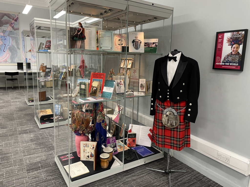 A mannequin dressed in jacket, sporran and kilt is placed beside a display of books, objects and artefacts in large glass cabinets.