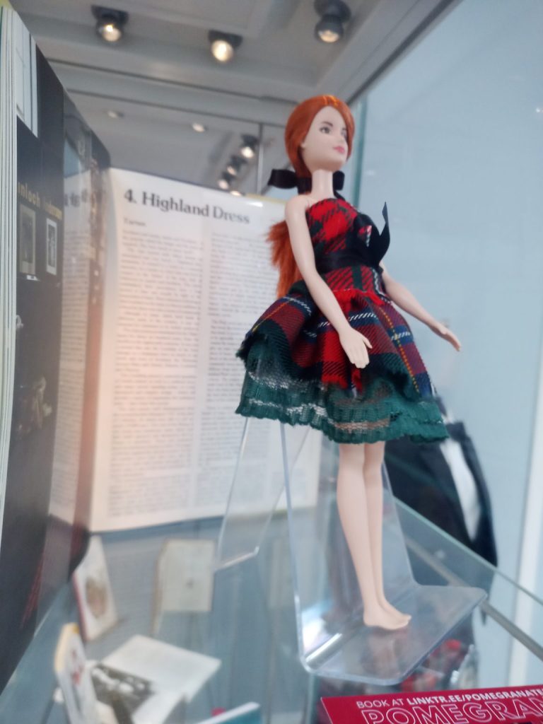 A Barbie doll wearing a tartan dress is displayed in a glass cabinet.